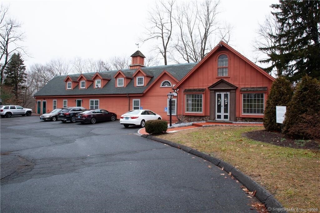 A two-story, red building with a cupola and multiple dormer windows, surrounded by parked cars and greenery.