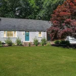 Charming Cape Cod-style home with a blue front door, white shutters, and a large, manicured lawn, surrounded by mature trees and a vibrant red-leafed tree in the front yard.