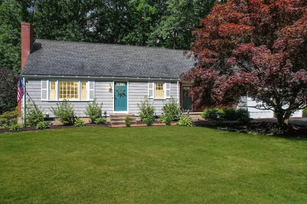 Charming Cape Cod-style home with a blue front door, white shutters, and a large, manicured lawn, surrounded by mature trees and a vibrant red-leafed tree in the front yard.