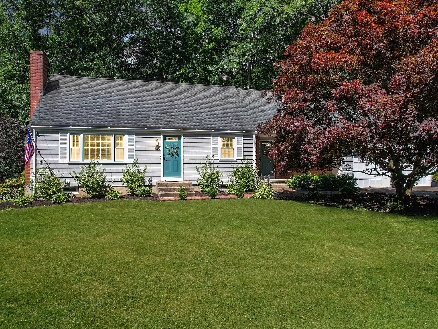 Charming Cape Cod-style home with a blue front door, white shutters, and a large, manicured lawn, surrounded by mature trees and a vibrant red-leafed tree in the front yard.