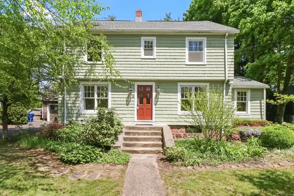 Front view of a charming two-story Colonial home with sage green siding, white trim, and a bright red front door, surrounded by lush greenery and mature trees