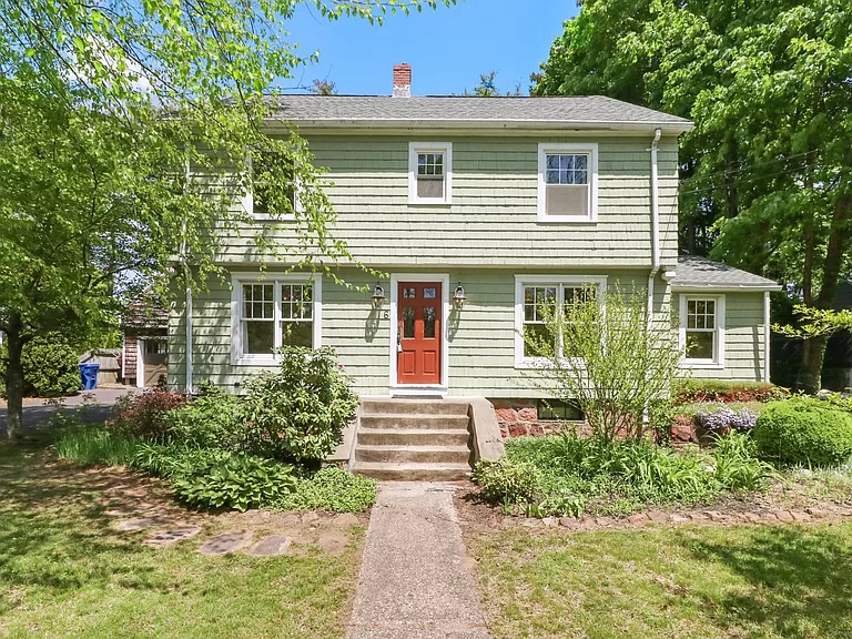 Front view of a charming two-story Colonial home with sage green siding, white trim, and a bright red front door, surrounded by lush greenery and mature trees