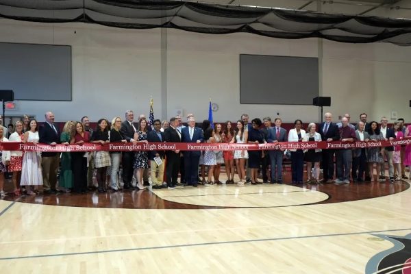 A large group of people, including officials and community members, stand together holding a long red ribbon with "Farmington High School" written on it during the ribbon-cutting ceremony inside a gymnasium