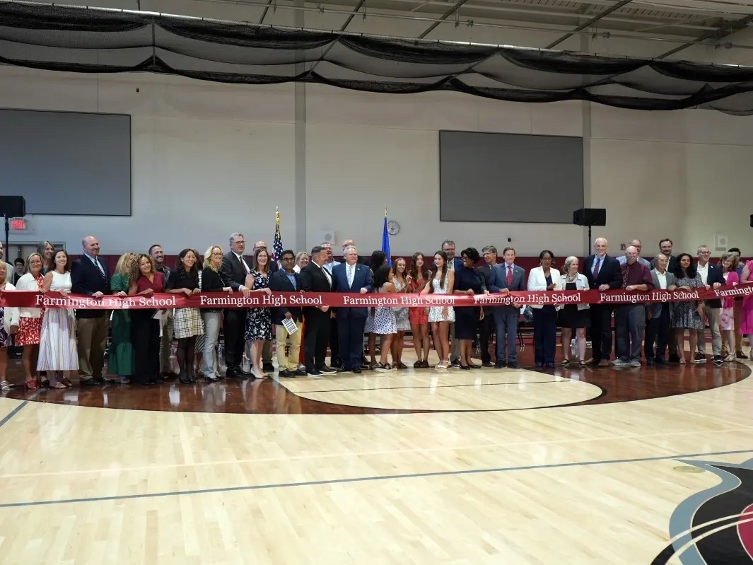 A large group of people, including officials and community members, stand together holding a long red ribbon with "Farmington High School" written on it during the ribbon-cutting ceremony inside a gymnasium