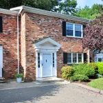 Front exterior of 6 Tunxis Village, Farmington, CT 06032, a red-brick townhouse with black shutters and a white entry door, surrounded by manicured shrubs and trees.