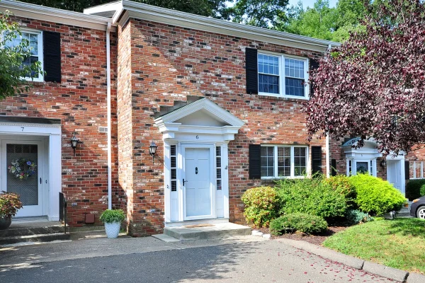 Front exterior of 6 Tunxis Village, Farmington, CT 06032, a red-brick townhouse with black shutters and a white entry door, surrounded by manicured shrubs and trees.