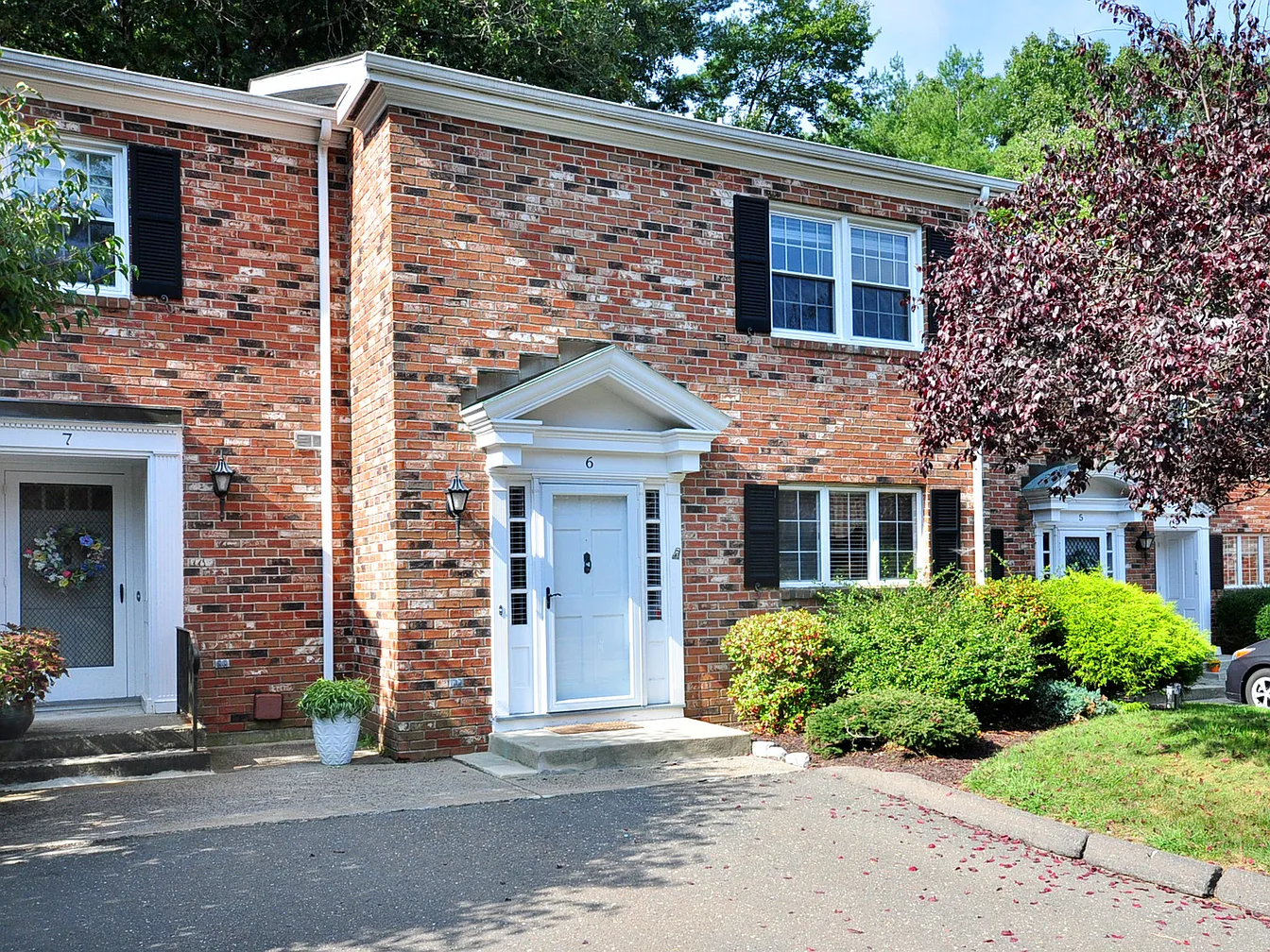 Front exterior of 6 Tunxis Village, Farmington, CT 06032, a red-brick townhouse with black shutters and a white entry door, surrounded by manicured shrubs and trees.