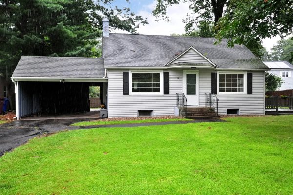 A cozy, gray Cape Cod-style home with white trim and black shutters at 12 Lake Garda Dr, Farmington, CT 06032. The house features a single-car carport, large windows, and a well-maintained lawn on a rainy day