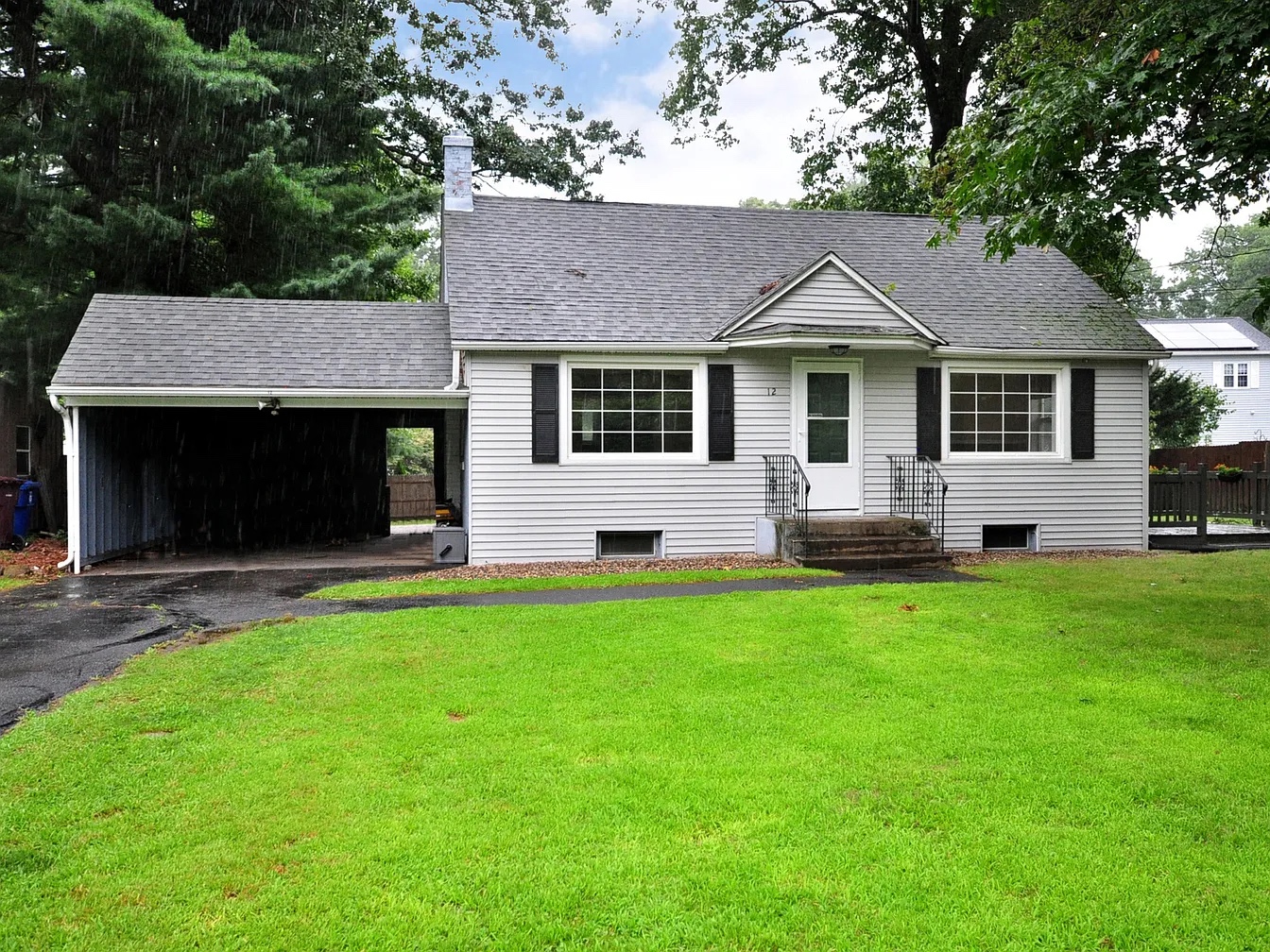 A cozy, gray Cape Cod-style home with white trim and black shutters at 12 Lake Garda Dr, Farmington, CT 06032. The house features a single-car carport, large windows, and a well-maintained lawn on a rainy day