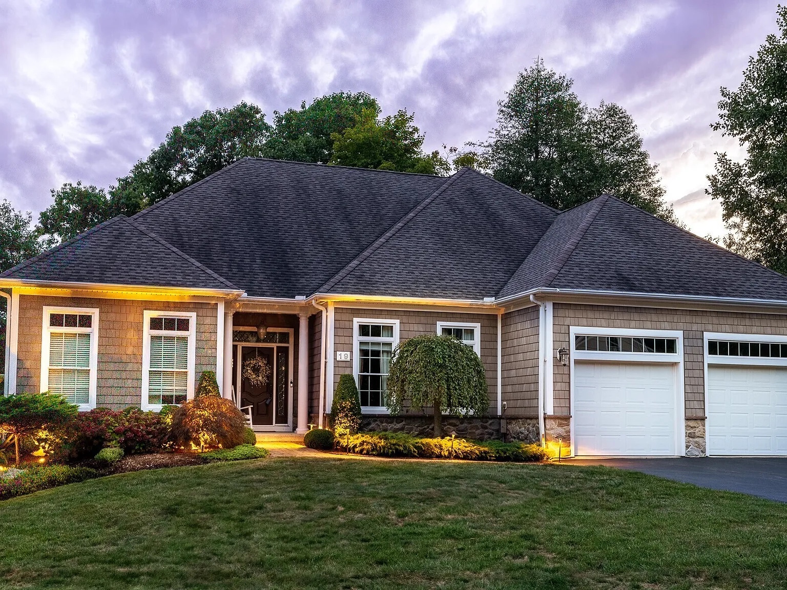 Exterior view of a luxury home at 19 Chimney Hill Dr, Farmington, CT 06032, featuring a two-car garage, well-manicured lawn, and warm exterior lighting at twilight.