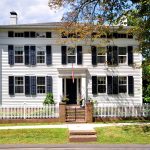 A historic white Colonial-style home with black shutters and a brick entryway, located at 16 Main St, Farmington, CT 06032, featuring a white picket fence and surrounded by greenery.