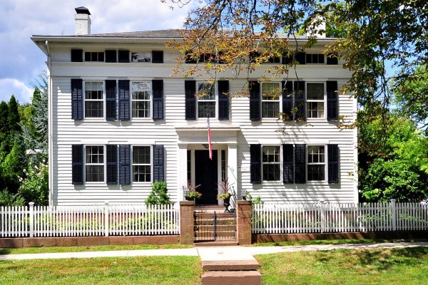 A historic white Colonial-style home with black shutters and a brick entryway, located at 16 Main St, Farmington, CT 06032, featuring a white picket fence and surrounded by greenery.