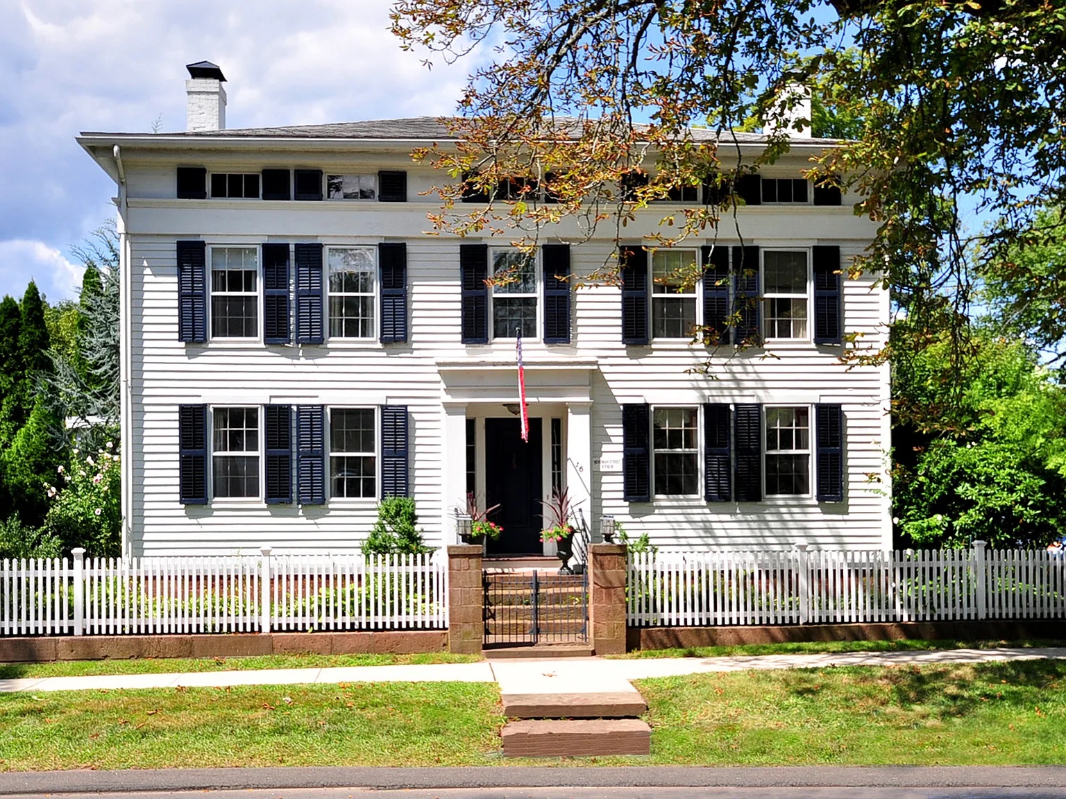 A historic white Colonial-style home with black shutters and a brick entryway, located at 16 Main St, Farmington, CT 06032, featuring a white picket fence and surrounded by greenery.