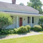 A charming single-story white house with a red front door, nestled in a lush green landscape, surrounded by bushes and trees. The home features black shutters and a brick walkway leading to the entrance.