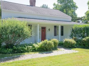 A charming single-story white house with a red front door, nestled in a lush green landscape, surrounded by bushes and trees. The home features black shutters and a brick walkway leading to the entrance.