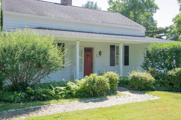 A charming single-story white house with a red front door, nestled in a lush green landscape, surrounded by bushes and trees. The home features black shutters and a brick walkway leading to the entrance.